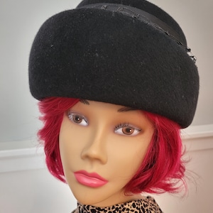 May include: A black hat with a rounded crown and a decorative ribbon detail. The hat is made of a felt-like material and is worn by a mannequin with red hair and a patterned scarf.