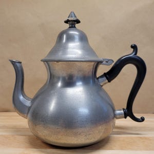 May include: A vintage pewter teapot with a black handle. The teapot has a bulbous body, a curved spout, and a lid with a decorative finial. The handle is curved and attached to the side of the pot. The teapot is sitting on a wooden surface.