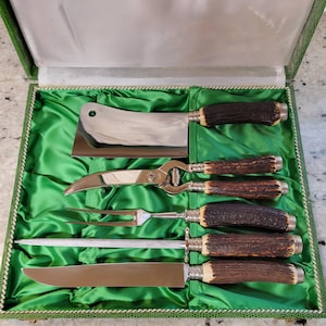 May include: A vintage carving set in a green lined case. The set includes a cleaver, shears, carving fork, sharpening steel, and carving knife. The handles are brown and silver, and the blades are shiny silver.