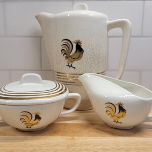 May include: A cream-colored ceramic tea set featuring a tall teapot with a lid, a lidded sugar bowl, and a creamer. Each piece is decorated with a gold and black rooster graphic and gold horizontal line accents. The teapot has a visible crackle glaze.