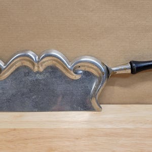 May include: A vintage silver-toned cheese knife with a decorative, wavy blade and a black wooden handle. The blade has a unique, scalloped design. The handle is curved for a comfortable grip. The knife is likely used for serving cheese.