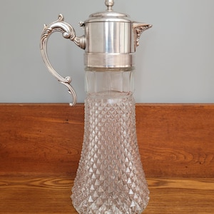 May include: A tall, clear glass pitcher with a silver-colored metal top, handle, and lid. The glass base has a diamond-patterned texture. The handle has an ornate, curved design. The pitcher is approximately 12 inches tall.