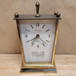 May include: Vintage Waltham clock with a white face, gold-tone frame, and decorative accents. The clock features a rectangular shape with a peaked top and a small, decorative finial. The clock face displays the word "JEWELED".
