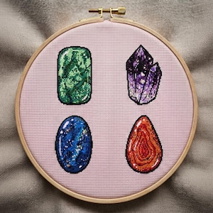 May include: A circular wooden embroidery hoop containing light pink fabric with four cross-stitched gemstone designs: a rectangular green stone, a pointed purple crystal cluster, an oval blue stone with white flecks, and a teardrop-shaped orange stone.
