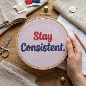 May include: A round wooden embroidery hoop containing light pink fabric with the cross-stitched text "Stay Consistent." The word "Stay" is stitched in red, and the word "Consistent." is stitched in dark blue, centered within the circular frame.