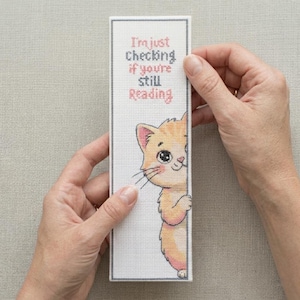 May include: A rectangular white bookmark featuring a cross-stitch design of a light orange kitten peeking from the side. The text reads, I'm just checking if you're still reading, in pink and gray lettering. The bookmark has a thin gray border.