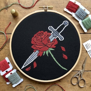 May include: Cross-stitch embroidery in a wooden hoop featuring a red rose pierced by a silver sword on a black background. Red petals fall from the rose. Embroidery floss, needles, and scissors surround the hoop on a wooden surface.