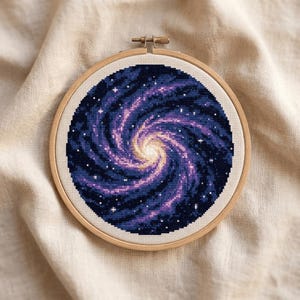 Puede incluir: Una pieza de punto de cruz circular con un diseño de galaxia espiral en tonos azul profundo, morado y amarillo brillante. La obra está asegurada dentro de un bastidor de madera redondo, mostrando un intrincado bordado de estrellas y polvo cósmico.