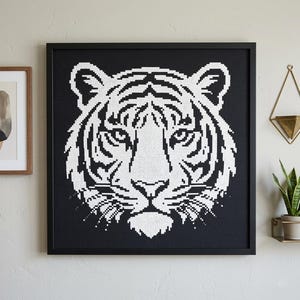 Puede incluir: Una obra de arte enmarcada en negro con un diseño de cara de tigre blanco. La cara del tigre está representada en un estilo pixelado sobre un fondo negro. La obra de arte cuadrada cuelga de una pared de color claro.