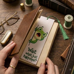 Puede incluir: Marcapáginas hecho a mano con un diseño de rana bordado a punto de cruz y el texto "Wait what just happened?". El marcapáginas está insertado en un libro encuadernado en cuero marrón. Hilo verde y borla adjuntos.