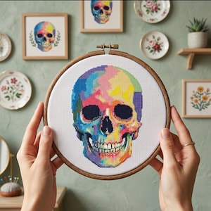 Puede incluir: Un bastidor de bordado circular de madera que contiene un panel de tela blanca con un diseño de punto de cruz de un cráneo humano. El cráneo está representado en una paleta de colores vibrantes que incluye tonos azules, amarillos, rosas y morados.