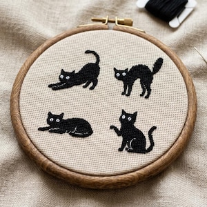 May include: A circular wooden embroidery hoop containing beige Aida fabric with four black cross-stitched cats in different poses. The cats feature white eyes and are arranged in a grid pattern. The hoop has a metal tightening screw at the top.