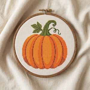 Puede incluir: Un bastidor de bordado circular de madera que contiene un diseño de punto de cruz de una calabaza naranja brillante con un tallo verde y una hoja verde. La calabaza presenta detalles acanalados verticales y un pequeño zarcillo rizado.