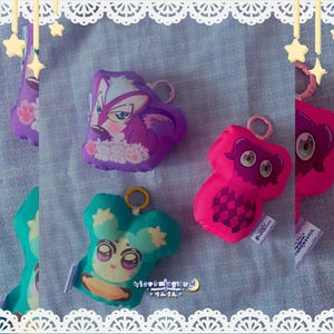 Puede incluir: Tres llaveros de peluche con personajes de dibujos animados. Uno es una criatura morada parecida a un zorro, otro es un monstruo rosa brillante con ojos grandes y patrón de cuadros, y otro es una criatura verde azulada con orejas.