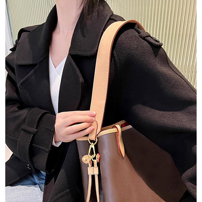 May include: A brown leather tote bag with a tan leather strap and gold hardware. The bag has a top zipper closure and a brown and tan patterned lining.