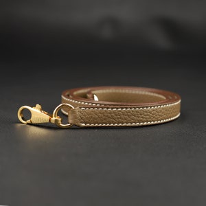 May include: A beige leather strap with a gold-toned metal clasp. The strap has a white stitch detail and is folded in half.