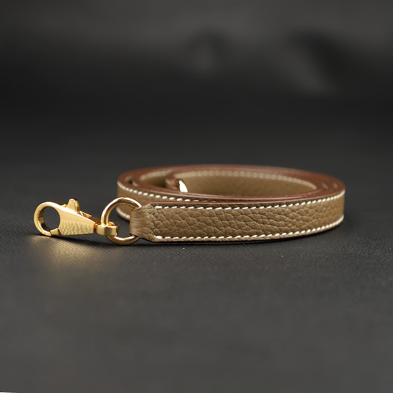 May include: A beige leather strap with a gold-toned metal clasp. The strap has a white stitch detail and is folded in half.