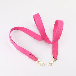 May include: A pink canvas strap with gold hardware and leather accents. The strap is designed for a bag or purse.