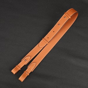 Peut inclure: Une sangle en cuir marron avec des coutures blanches et des accents argent&eacute;s. La sangle est pli&eacute;e en deux et pos&eacute;e &agrave; plat sur une surface noire.