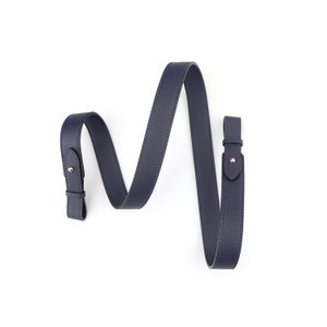 Puede incluir: Una correa de cuero azul oscuro con herrajes dorados. La correa está enrollada y lista para ser fijada a un bolso u otro artículo.