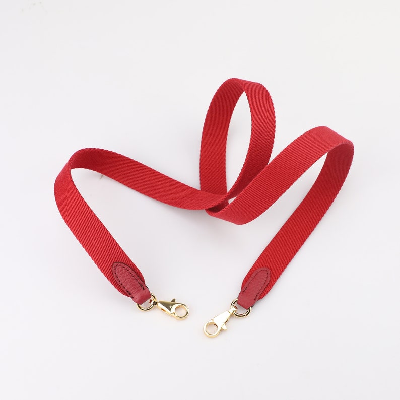 May include: Red canvas shoulder strap with gold metal clasps. The strap is designed for a handbag or purse.
