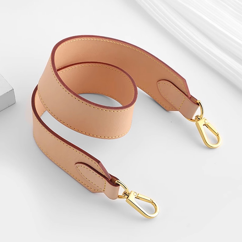 May include: A light brown leather shoulder strap with gold metal clasps. The strap is folded in half and the clasps are visible.