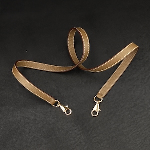 May include: A light brown leather strap with gold-toned metal clasps. The strap is approximately 45 inches long and 1 inch wide.