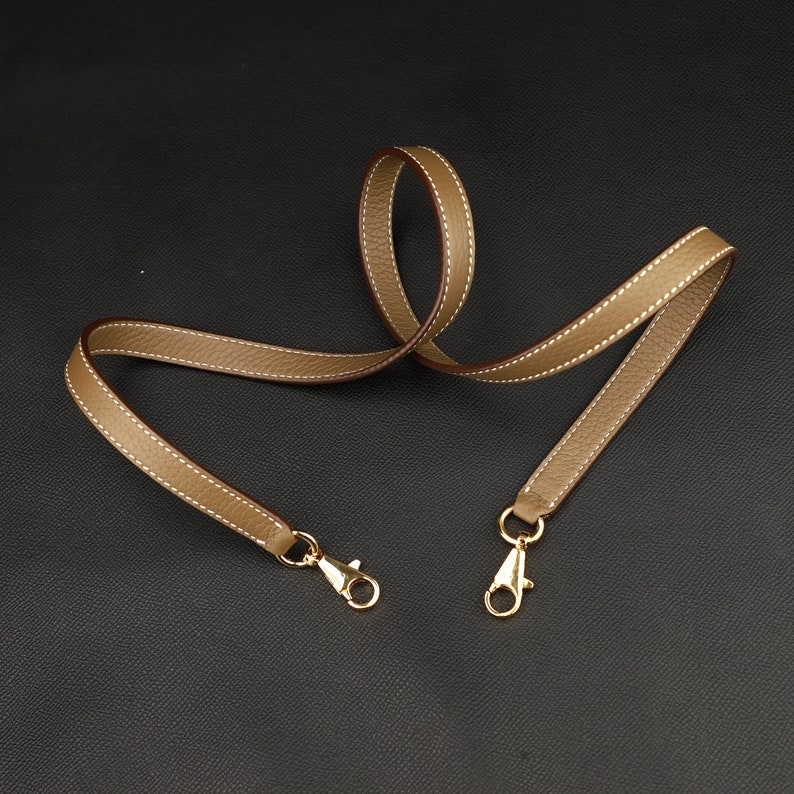 May include: A light brown leather strap with gold-toned metal clasps. The strap is approximately 45 inches long and 1 inch wide.