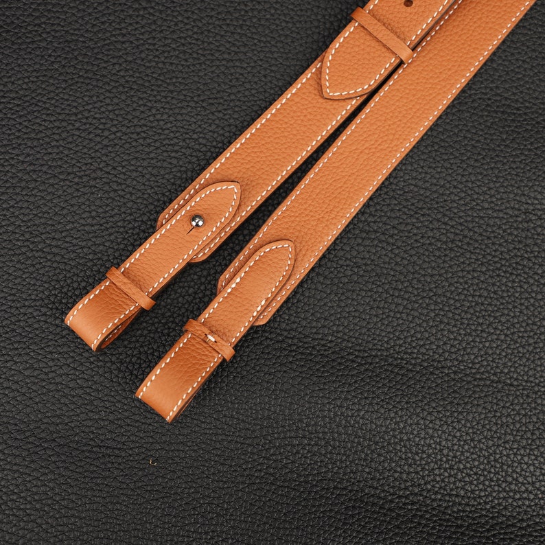 Peut inclure: Deux lani&egrave;res en cuir marron avec des coutures blanches. Les lani&egrave;res sont sur un fond textur&eacute; noir.