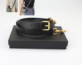 Könnte beinhalten: Zwei schwarze Lederriemen mit goldenen Beschlägen, darunter eine Schnalle und ein Haken. Die Riemen liegen auf einer schwarzen Box.