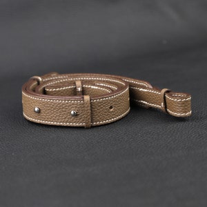 Peut inclure: Une bandouli&egrave;re d'appareil photo en cuir marron avec des coutures blanches et des rivets argent&eacute;s. La bandouli&egrave;re est enroul&eacute;e et poss&egrave;de une petite boucle &agrave; une extr&eacute;mit&eacute;.