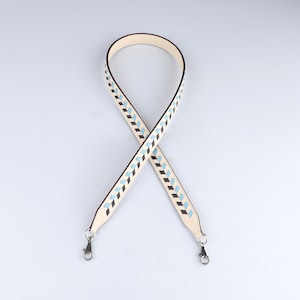 May include: A cream-colored bag strap featuring a repeating geometric chevron pattern in light blue and dark brown. The strap is constructed with a smooth finish and includes silver-toned metal lobster claw clasps at both ends for attachment.