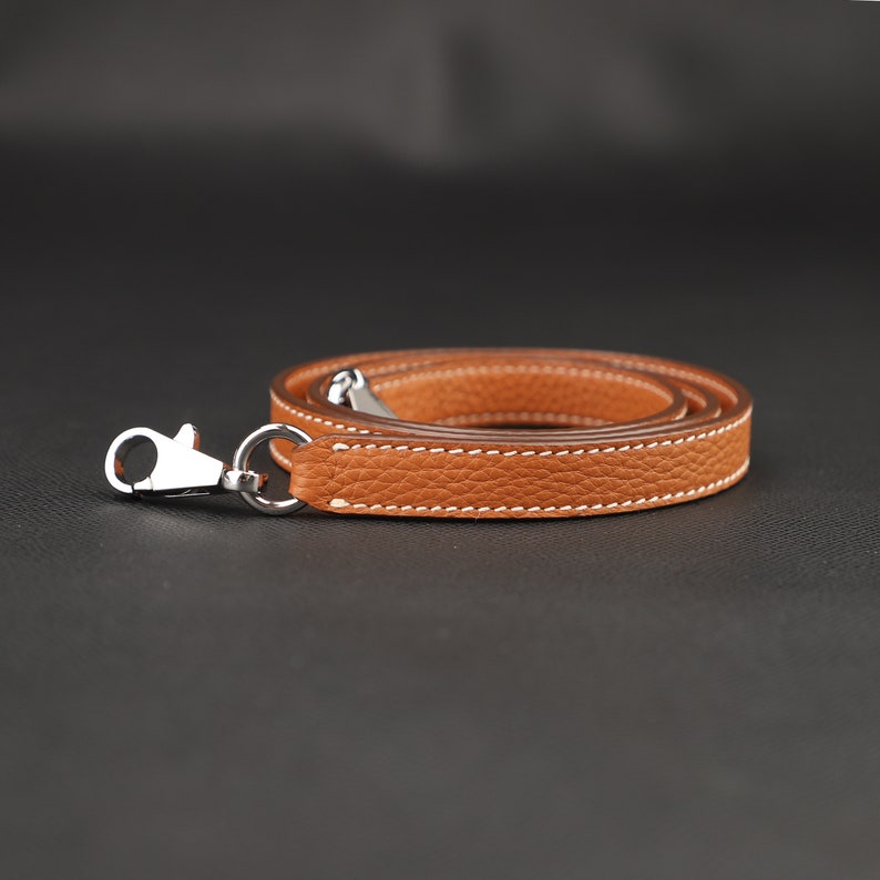 May include: A brown leather strap with a silver hook. The strap is folded in half and the hook is at the end of the strap.
