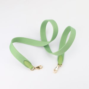 May include: A light green canvas strap with gold metal clasps and leather accents. The strap is designed for a bag or purse.