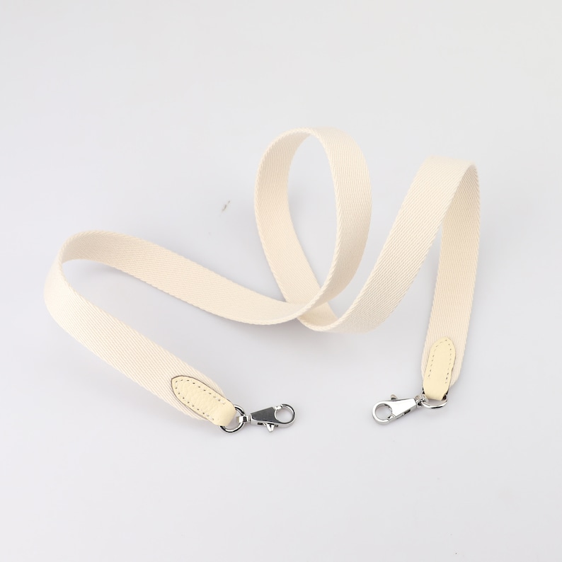 May include: A cream-colored canvas shoulder strap with two silver clasps and two light brown leather tabs.