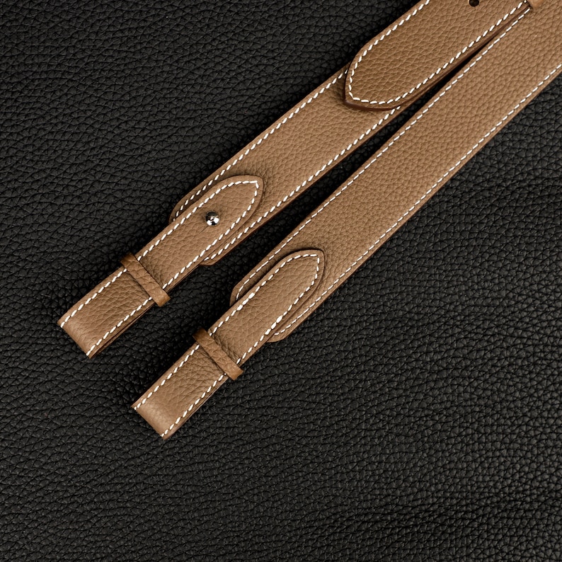 Peut inclure: Deux lani&egrave;res en cuir marron avec des coutures blanches. Les lani&egrave;res sont dispos&eacute;es sur une surface textur&eacute;e noire.