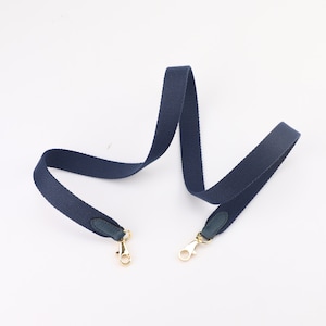 May include: A dark blue canvas strap with gold clasps and dark blue leather accents. The strap is designed for a bag or purse.