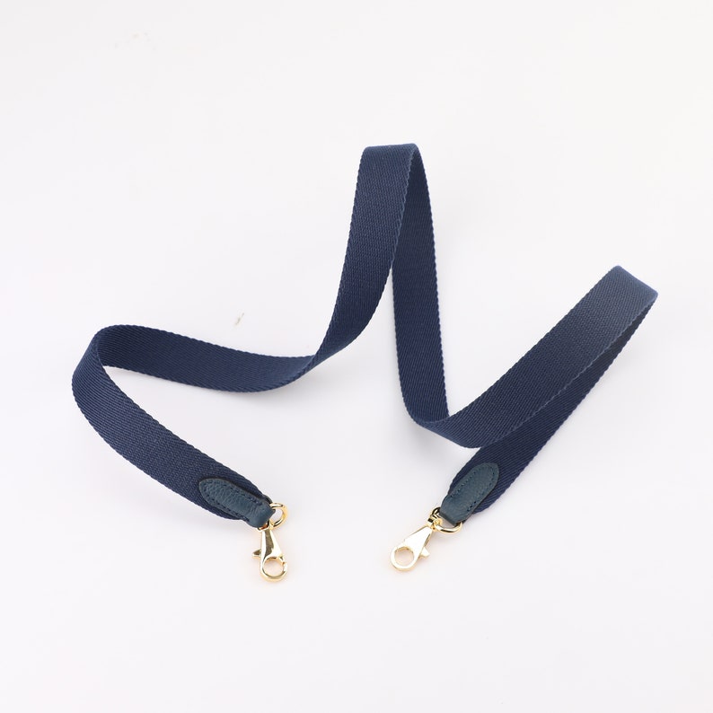 May include: A dark blue canvas strap with gold clasps and dark blue leather accents. The strap is designed for a bag or purse.