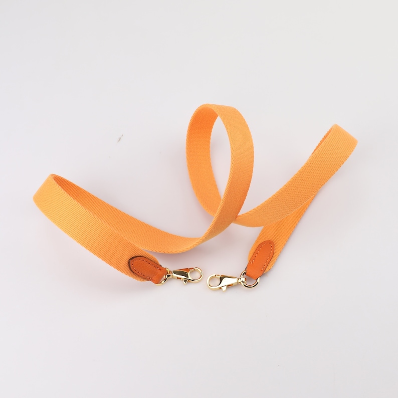 May include: A bright orange canvas strap with brown leather accents and gold metal clasps. The strap is designed for a bag or purse.