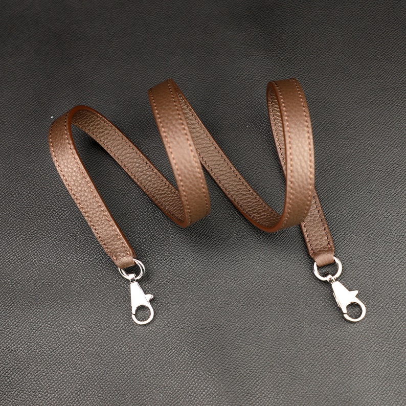 May include: A brown leather strap with silver clasps. The strap is coiled and ready to be attached to a bag or other item.
