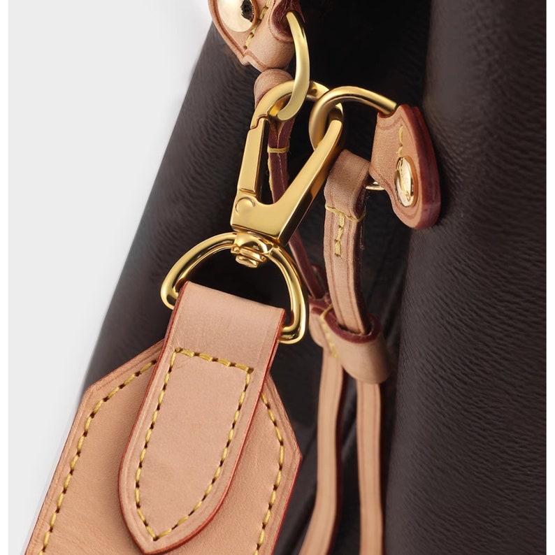 May include: Close-up of a gold-toned metal clasp attached to a brown leather strap with a light brown leather patch. The strap has yellow stitching.