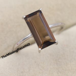 May include: A silver ring featuring a rectangular, smoky brown gemstone. The gemstone is set with four prongs, and the band is a simple, polished silver. The ring is displayed on a soft, textured surface. The text "TheMissingPieceIND" is visible.