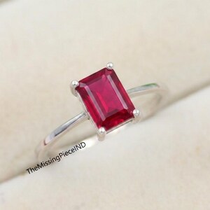 Peut inclure: Une bague en argent ornée d'une pierre précieuse rectangulaire rouge foncé. La pierre est le point central, sertie dans un anneau simple et élégant. La bague est présentée sur une surface douce et claire, soulignant son design.