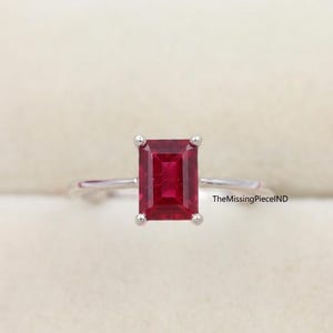May include: A silver ring with a rectangular, deep red gemstone. The gemstone is the focal point, held in place by four prongs. The ring's band is thin and polished. The text "TheMissingPieceIND" is visible on the right side.