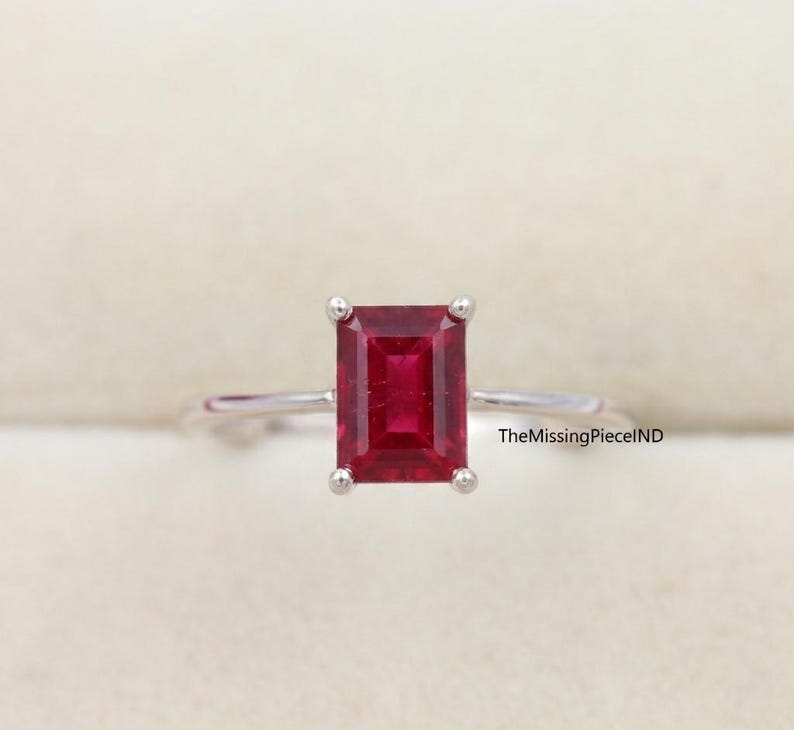 May include: A silver ring with a rectangular, deep red gemstone. The gemstone is the focal point, held in place by four prongs. The ring's band is thin and polished. The text "TheMissingPieceIND" is visible on the right side.