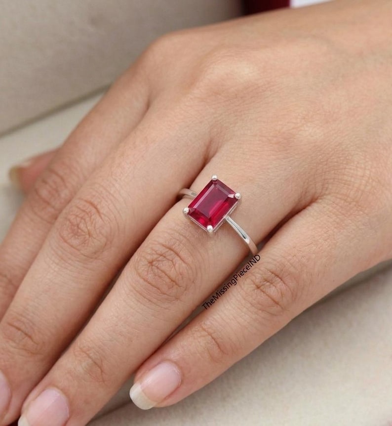 May include: A silver ring with a rectangular, deep red gemstone. The ring is set on a slender band, highlighting the vibrant colour of the stone. The MissingPieceIND is visible on the side of the ring.