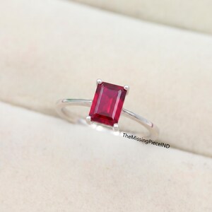 May include: A silver ring with a rectangular, deep red gemstone. The gemstone is the focal point, held in place by four prongs. The ring's band is thin and polished, creating a minimalist design. The ring is displayed on a neutral background.