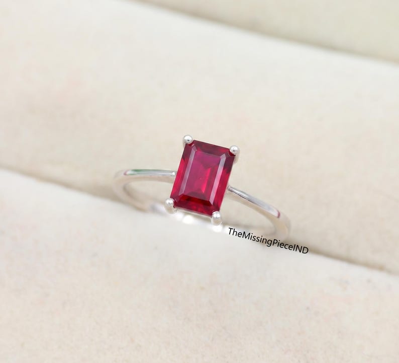 May include: A silver ring with a rectangular, deep red gemstone. The gemstone is the focal point, held in place by four prongs. The ring's band is thin and polished, creating a minimalist design. The ring is displayed on a neutral background.