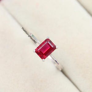 May include: A silver ring with a rectangular, deep red gemstone. The gemstone is held in place by four prongs. The ring band is slender and shiny. TheMissingPieceND is printed on the ring band.