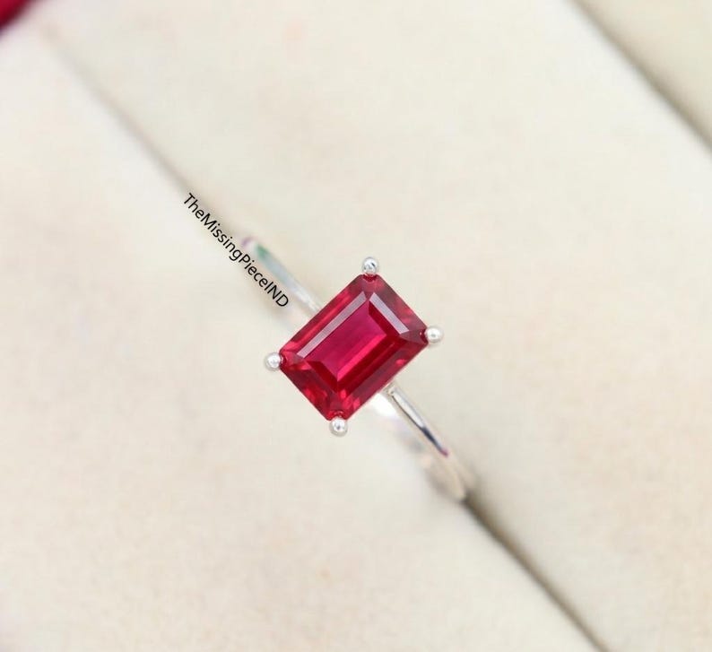 May include: A silver ring with a rectangular, deep red gemstone. The gemstone is held in place by four prongs. The ring band is slender and shiny. TheMissingPieceND is printed on the ring band.
